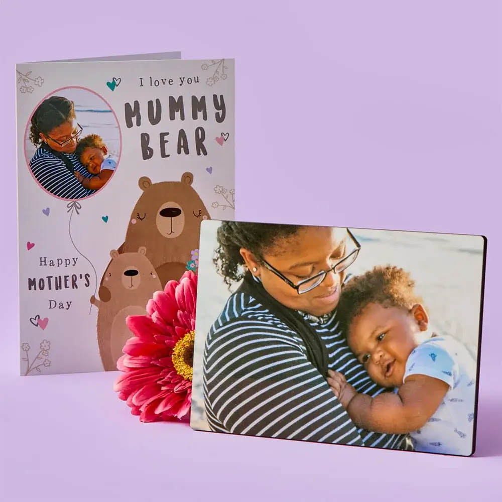 A Mother’s Day card with illustrated bears stands behind a photo plaque showing a parent holding a child, with a bright pink flower placed in front on a pastel purple background.